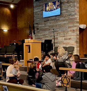 A group of kids sit with Tricia Grafelman on the steps at the front of the sanctuary for the Young Disciples segment of worship service.