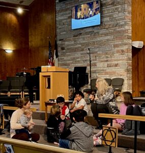 A group of kids sit with Tricia Grafelman on the steps at the front of the sanctuary for the Young Disciples segment of worship service. 